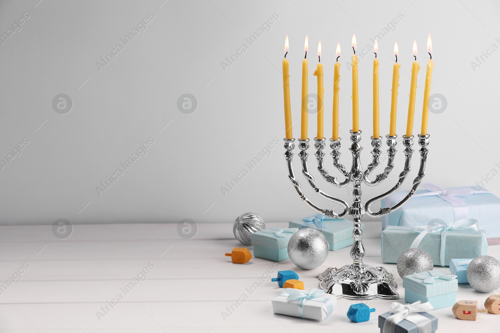 Photo of Hanukkah celebration. Menorah with burning candles, dreidels, gift boxes and holiday ornaments on white wooden table, space for text