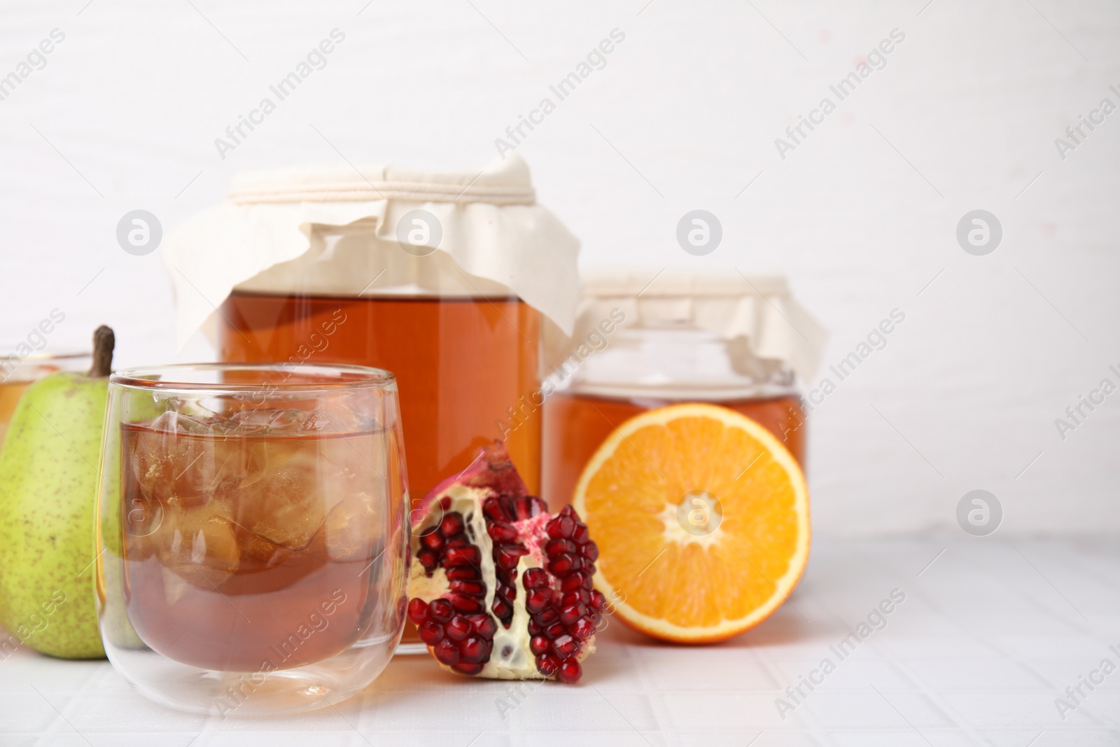 Photo of Tasty kombucha with ice cubes and fresh fruits on white tiled table, space for text