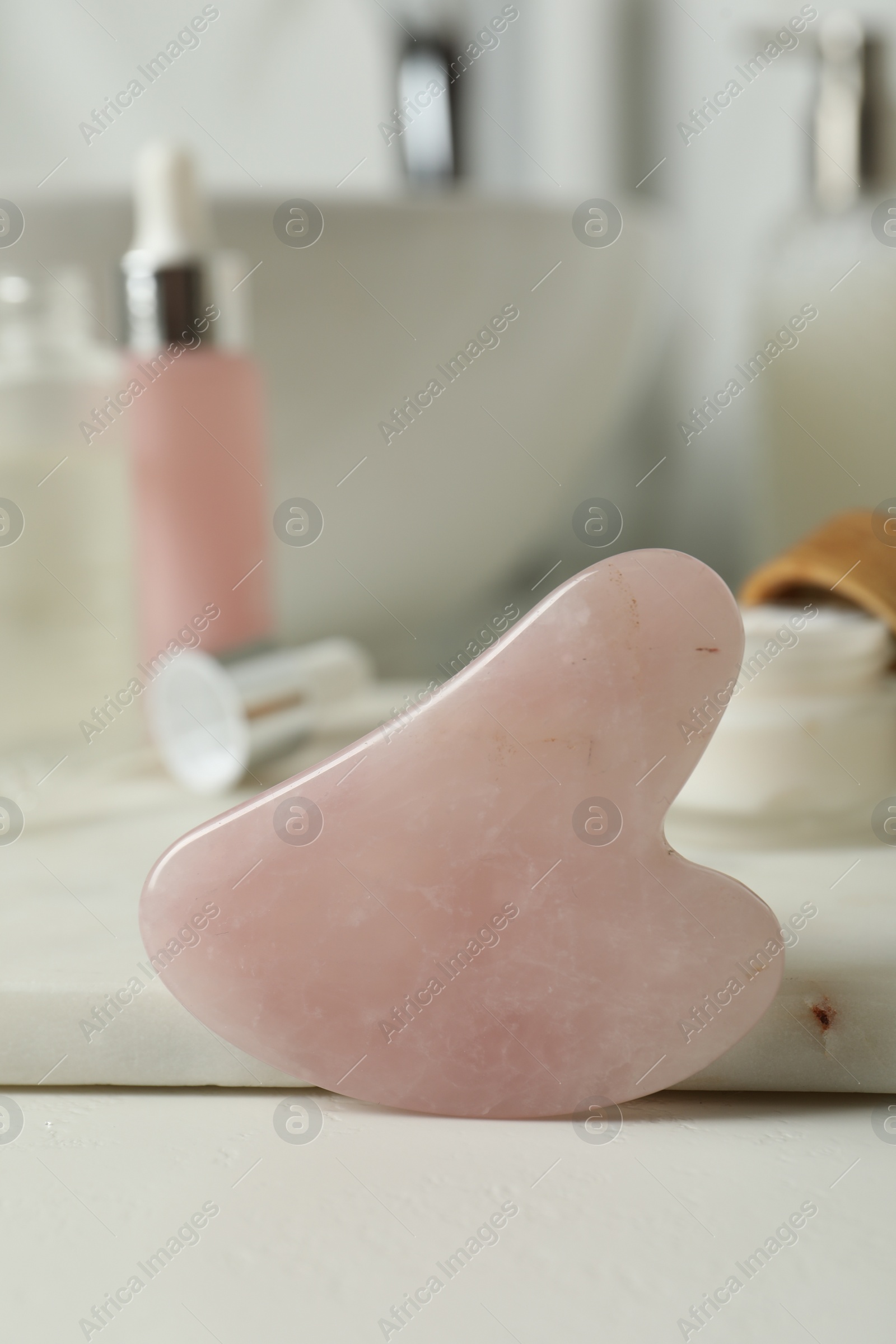 Photo of Rose quartz gua sha tool and toiletries on white countertop in bathroom, closeup