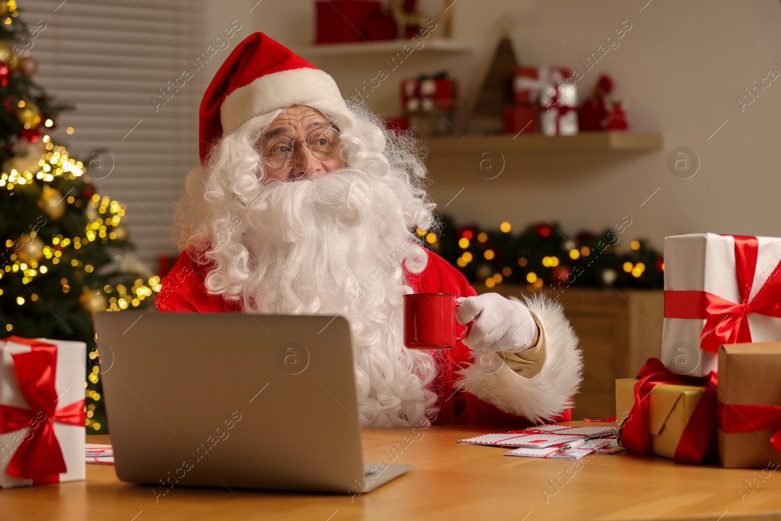 Photo of Santa Claus holding cup of drink using laptop at table with Christmas gifts in room