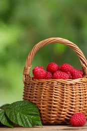 Photo of Wicker basket with tasty ripe raspberries and green leaves on table against blurred background, closeup