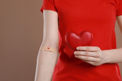 Photo of Blood donation concept. Woman with adhesive plaster on arm holding red heart against brown background, closeup. Space for text