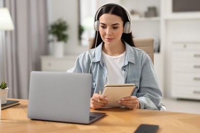 Photo of Young woman in headphones watching webinar at table in room