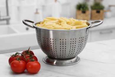 Cooked pasta in metal colander and tomatoes on light marble table, closeup