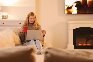 Young woman with cup of hot drink working on laptop near fireplace at home
