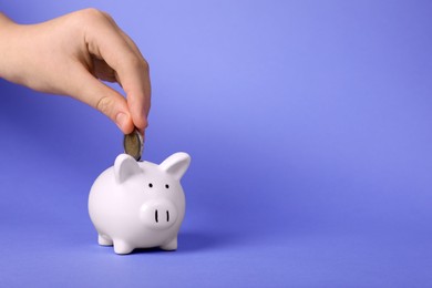 Photo of Woman putting coin into ceramic piggy bank on purple background, closeup with space for text. Financial savings