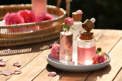 Bottles of rose essential oil and flowers on wooden table outdoors, space for text