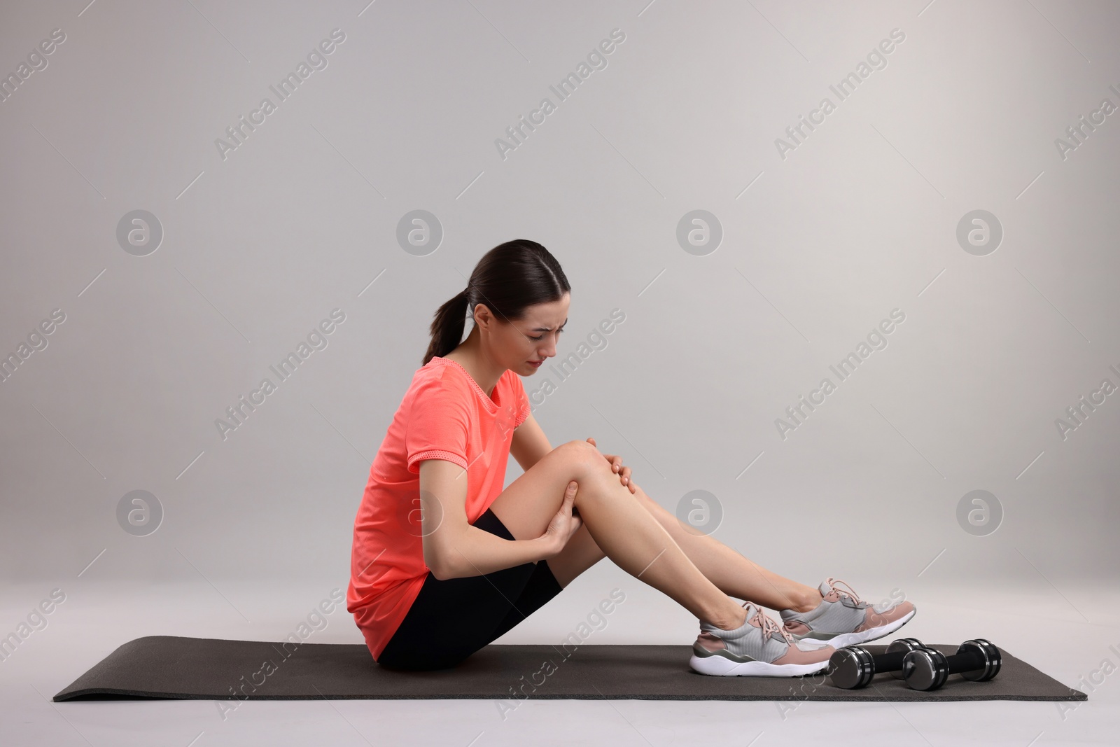 Photo of Young woman suffering from leg pain on exercise mat against grey background
