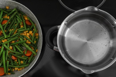 Pot near frying pan with vegetables on electric stove, top view