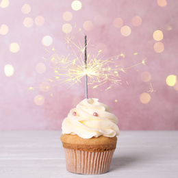 Image of Birthday cupcake with sparkler on white wooden table against light pink background