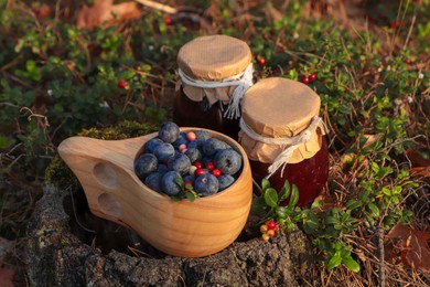 Photo of Wooden mug full of fresh ripe blueberries, lingonberries and jars with jam on stump