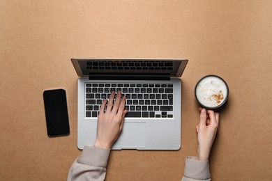 Woman with cup of cappuccino using laptop at light brown table, top view