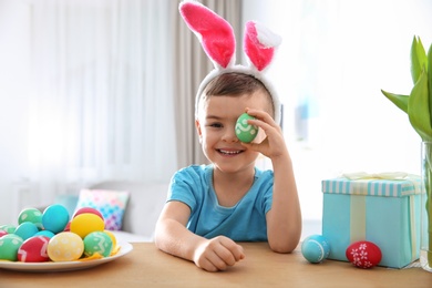 Photo of Cute little boy with bunny ears headband playing with painted Easter egg in room