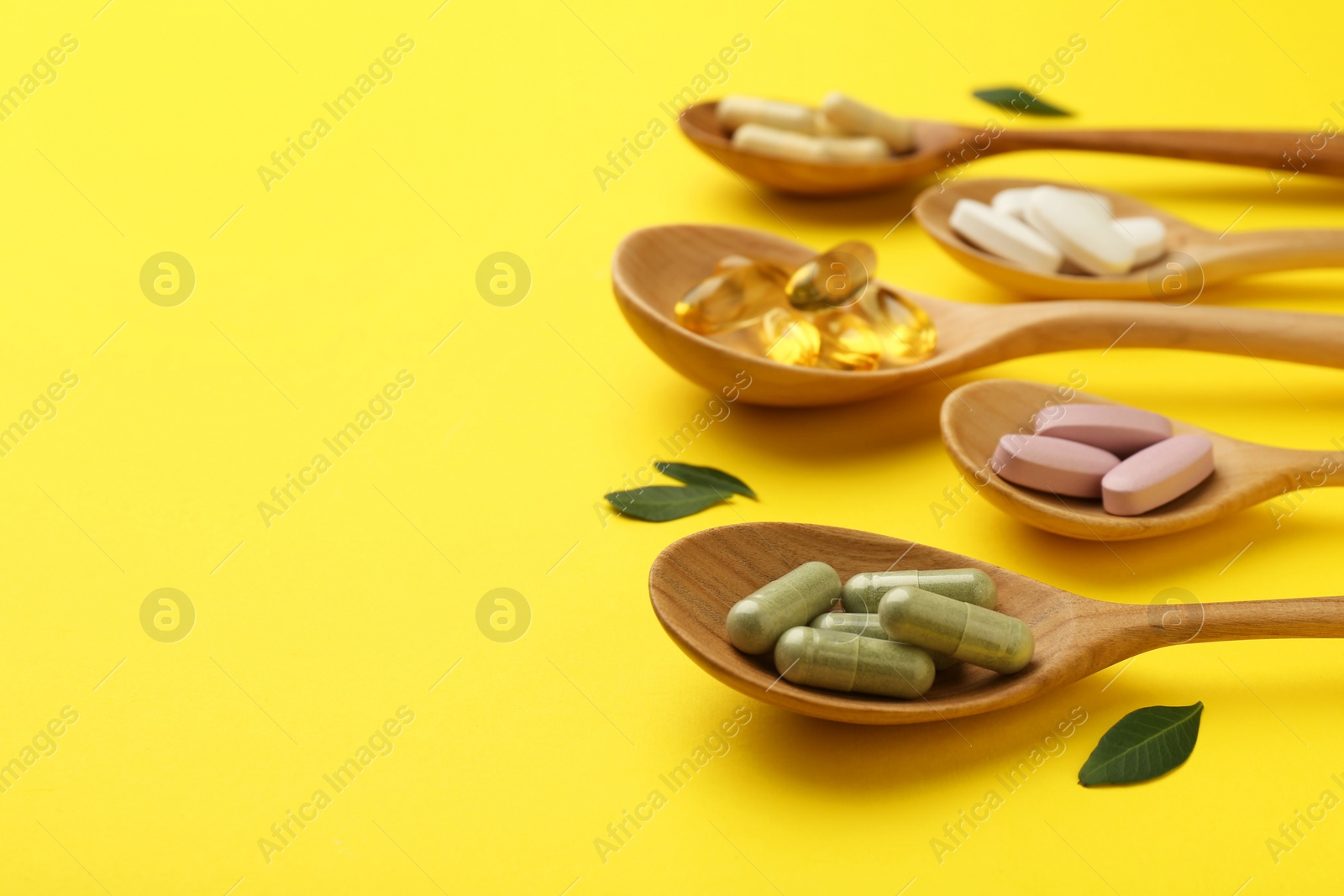 Photo of Different vitamin pills in spoons and green leaves on yellow background, closeup. Space for text