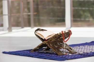 Photo of Rehal with open Quran and Misbaha on Muslim prayer rug near window indoors, space for text