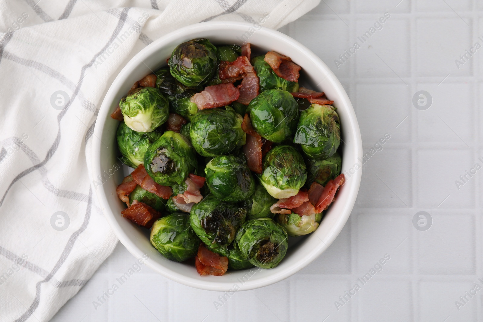 Photo of Delicious roasted Brussels sprouts and bacon in bowl on white tiled table, top view