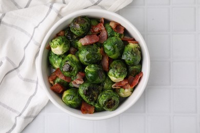 Delicious roasted Brussels sprouts and bacon in bowl on white tiled table, top view