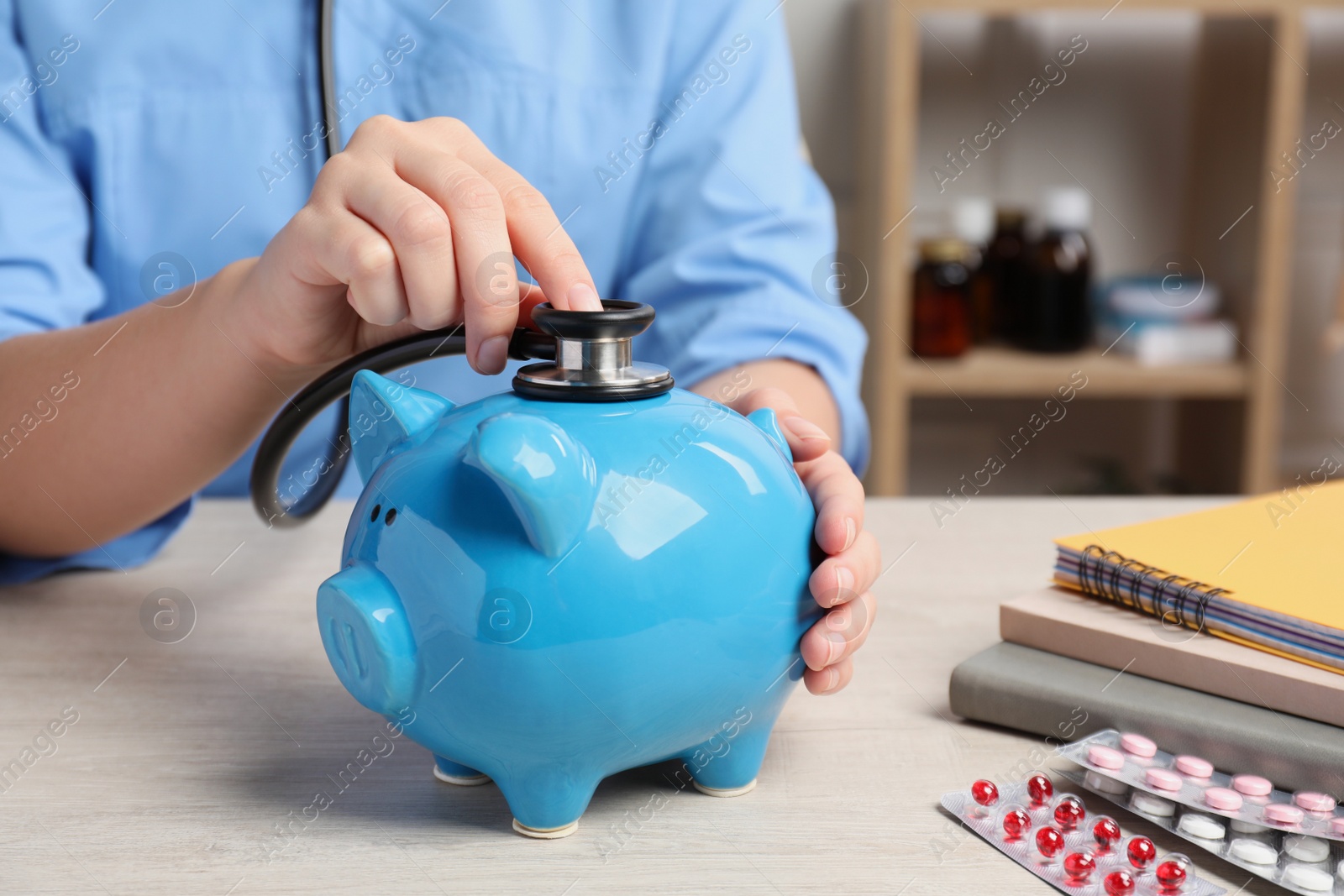 Photo of Doctor with stethoscope and piggy bank near pills at wooden table in hospital, closeup. Medical insurance