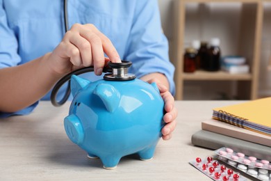 Doctor with stethoscope and piggy bank near pills at wooden table in hospital, closeup. Medical insurance