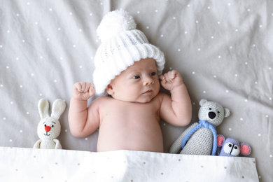 Cute newborn baby wearing white knitted hat with toys in bed, top view