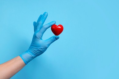 Photo of Doctor wearing medical glove holding decorative heart on light blue background, closeup. Space for text