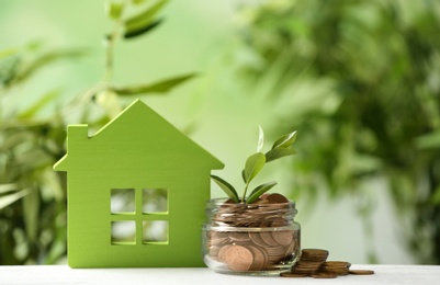 Model of house near jar with coins and plant on table against blurred white background. Space for text
