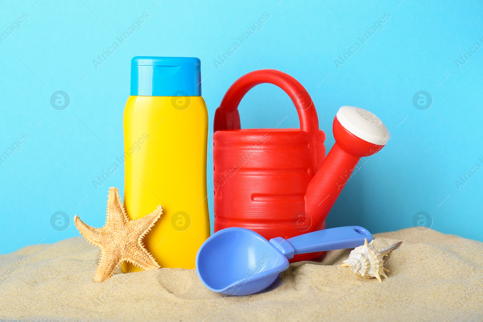 Photo of Suntan product, starfish and plastic beach toys on sand against light blue background