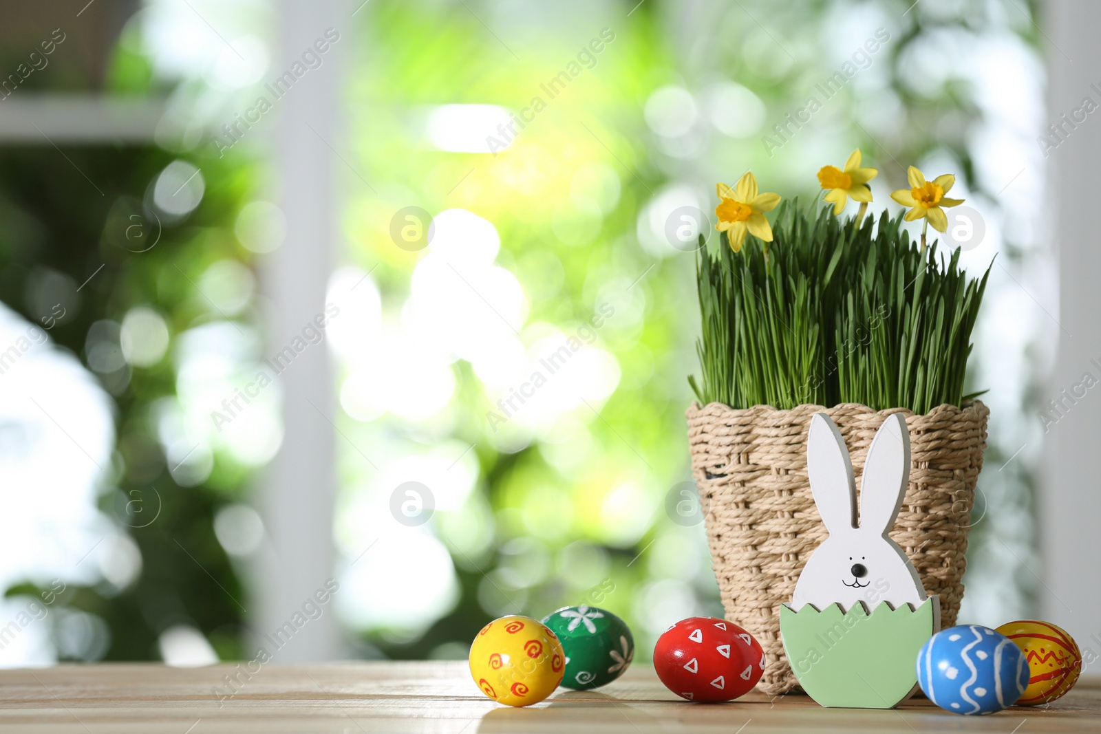 Photo of Easter bunny figure, dyed eggs and flowers on wooden table against blurred green background. Space for text