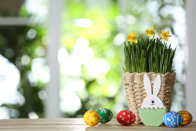 Photo of Easter bunny figure, dyed eggs and flowers on wooden table against blurred green background. Space for text