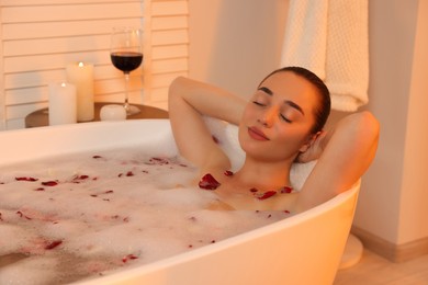 Photo of Woman taking bath in tub with foam and rose petals indoors