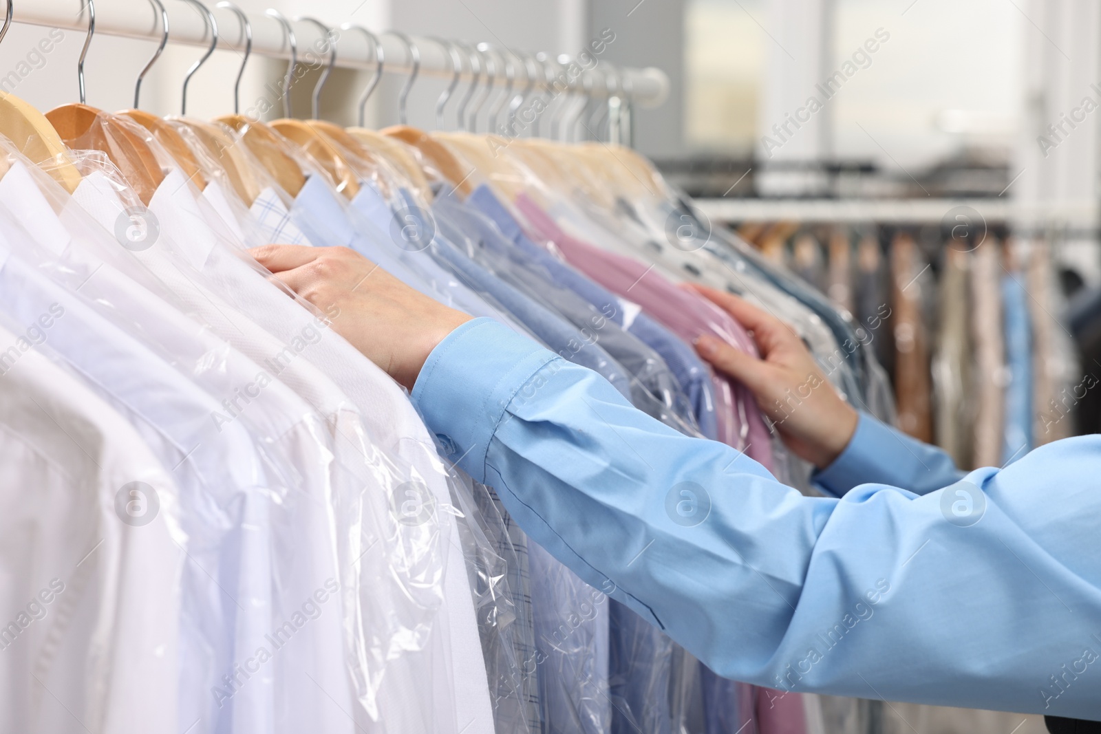 Photo of Dry-cleaning service. Woman taking shirt in plastic bag from rack indoors, closeup