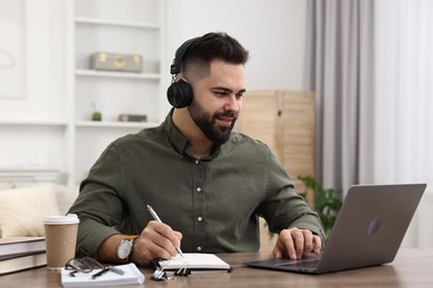 E-learning. Young man taking notes during online lesson at wooden table indoors