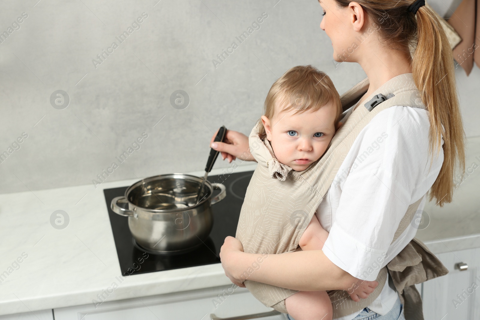 Photo of Mother holding her child in sling (baby carrier) while cooking in kitchen