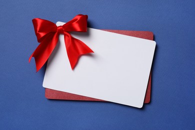 Photo of Blank gift card with red bow on blue background, top view. Space for text