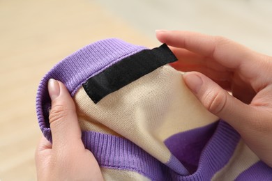 Photo of Woman holding colorful sweater with blank clothing label on blurred background, closeup