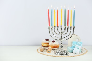 Photo of Hanukkah celebration. Menorah with burning candles, dreidels, donuts and gift boxes on white wooden table, space for text