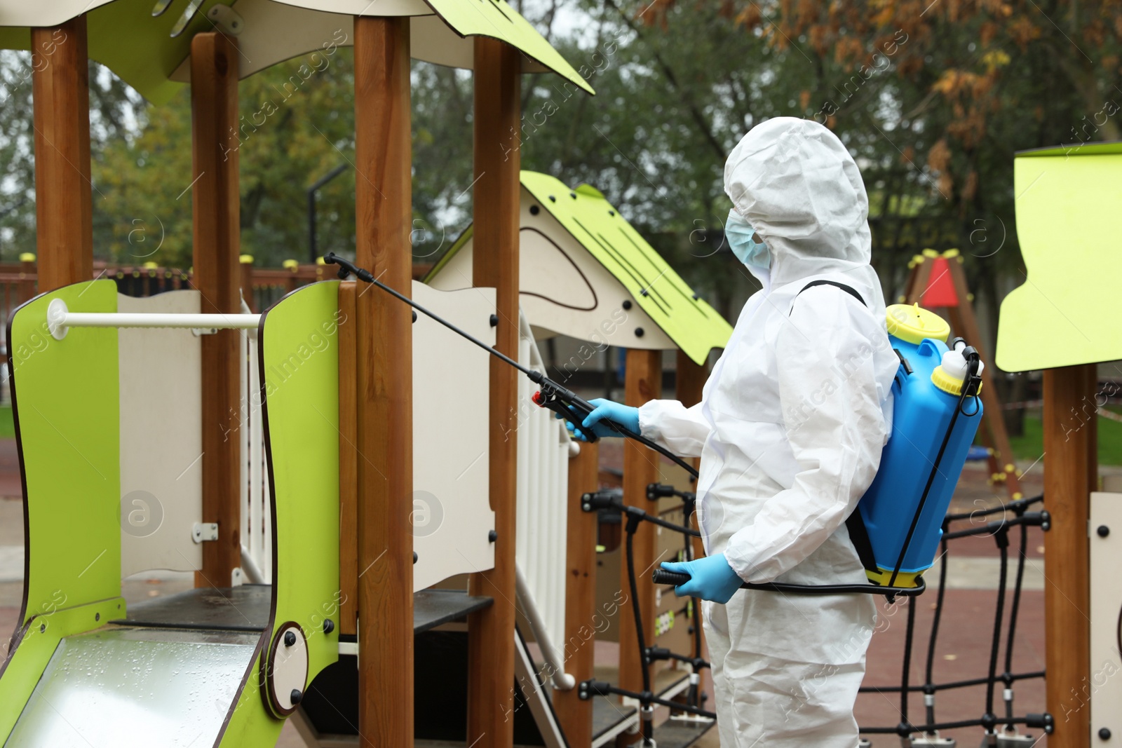 Photo of Woman wearing chemical protective suit with disinfectant sprayer on playground. Preventive measure during coronavirus pandemic