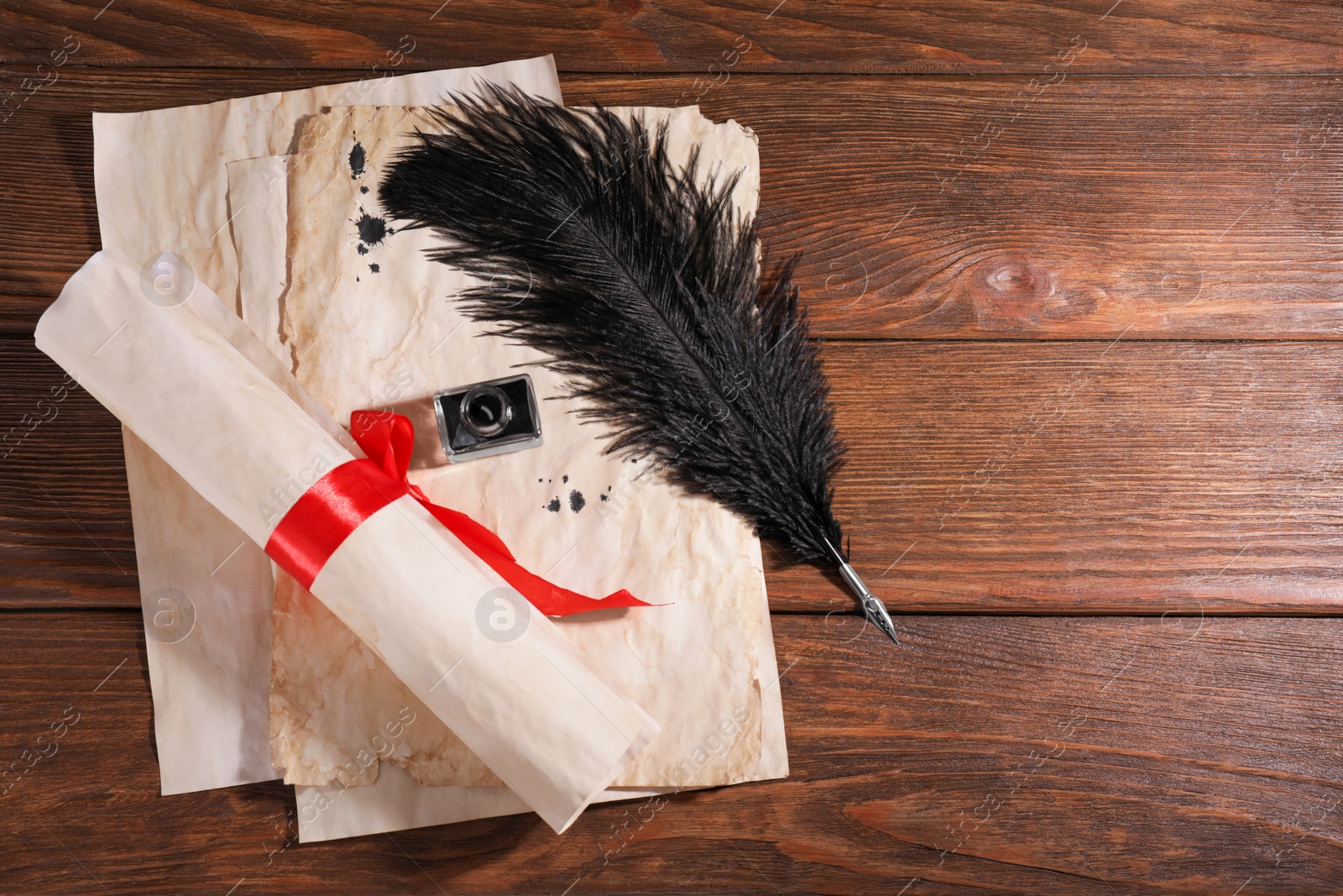 Photo of Black feather pen, inkwell and vintage parchment with ink stains on wooden table, flat lay. Space for text