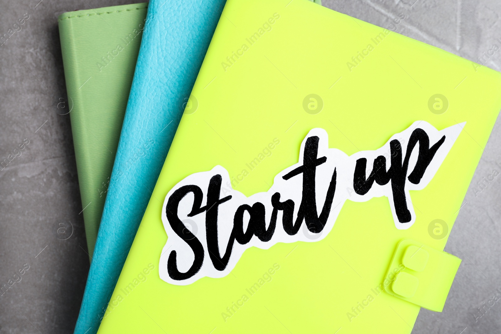 Photo of Sheet of paper with phrase Start Up and notebooks on grey table, top view