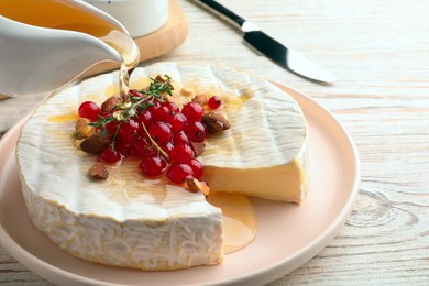Photo of Pouring honey onto brie cheese served with almonds and red currants on white wooden table, closeup