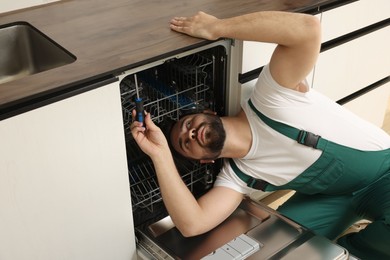 Photo of Serviceman repairing dishwasher with screwdriver in kitchen