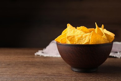 Photo of Tortilla chips (nachos) in bowl on wooden table against dark background. Space for text