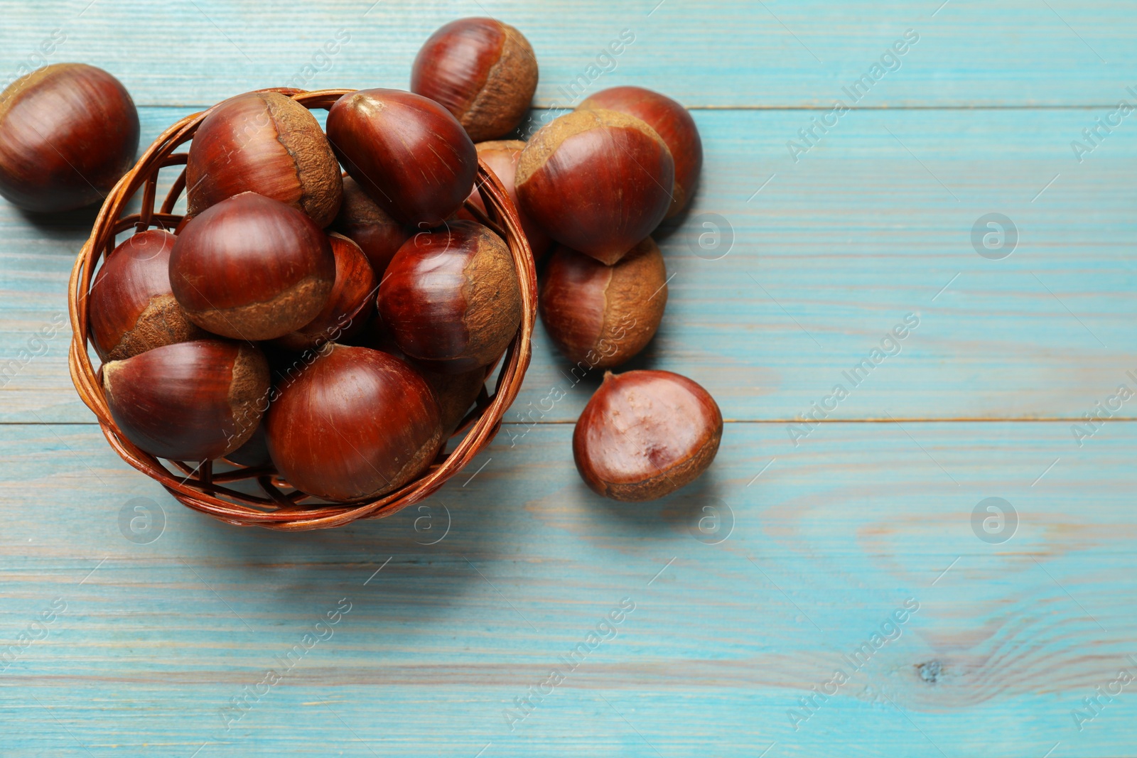 Photo of Wicker bowl with roasted edible sweet chestnuts on light blue wooden table, flat lay. Space for text