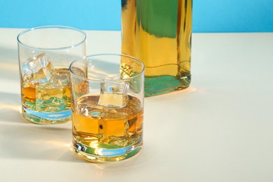 Whiskey with ice cubes in glasses and bottle on white table, space for text