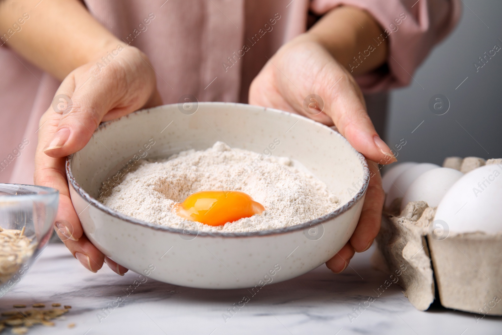 Photo of Woman holding ceramic bowl with flour and egg at white marble table in kitchen, closeup. Cooking oatmeal dough