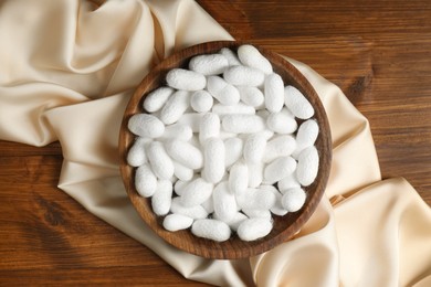Photo of White cocoons with bowl and silk fabric on wooden table, flat lay