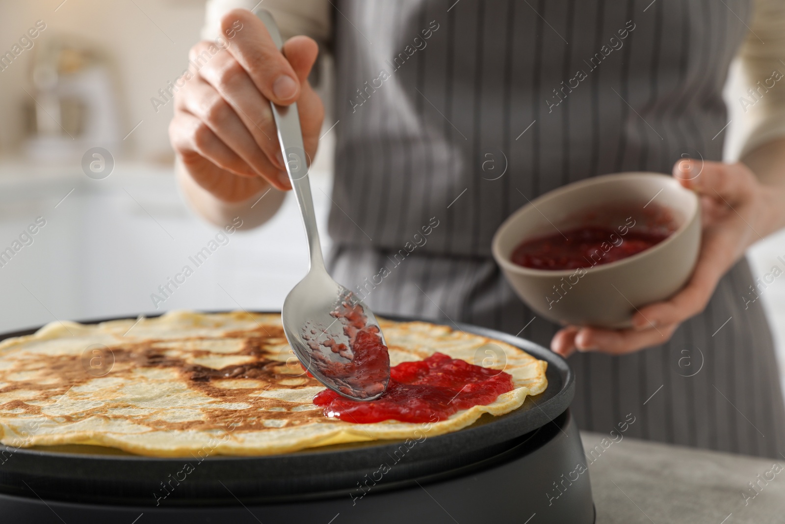 Photo of Woman cooking delicious crepe with jam on electric pancake maker in kitchen, closeup