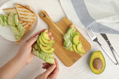 Woman holding tasty avocado toast over white wooden table, top view