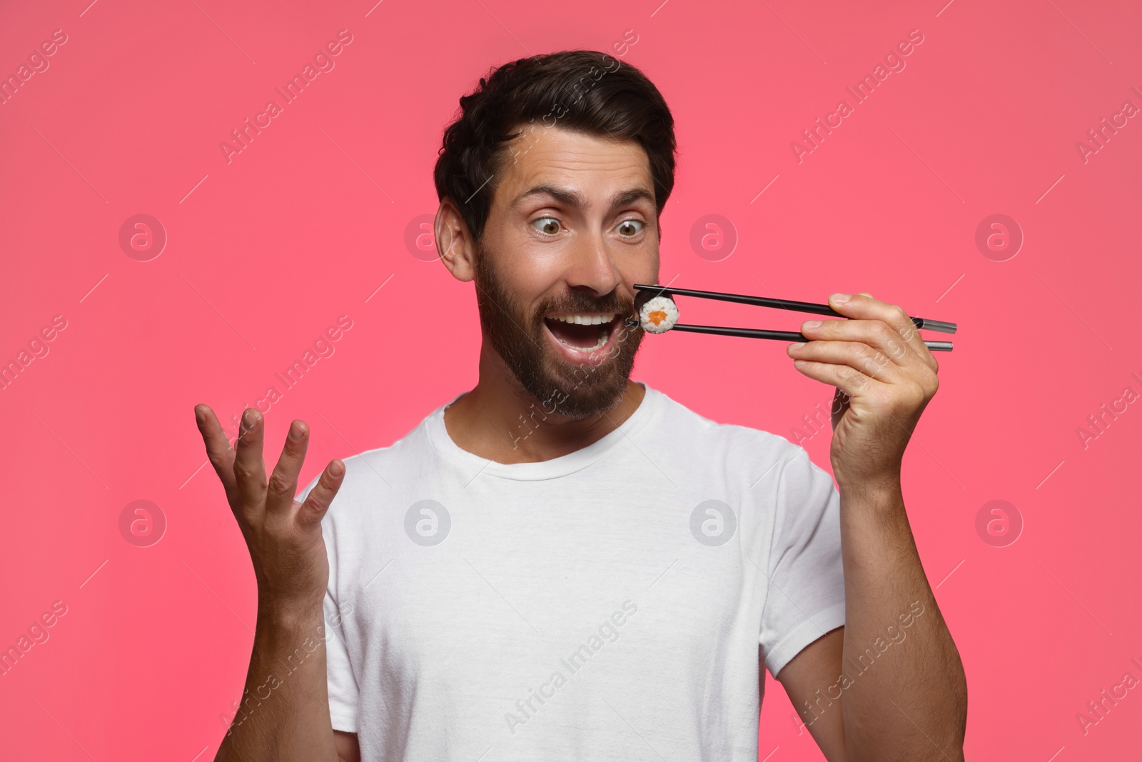 Photo of Emotional man holding tasty sushi roll with chopsticks on pink background