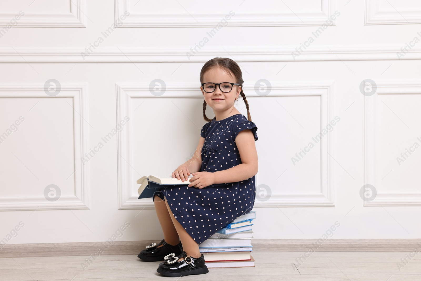 Photo of Cute little girl in glasses sitting on stack of books near white wall